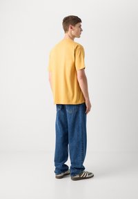 Gul bomull t-skjorte med avslappet passform, korte ermer og lette sidespalter. Kombinert med løse blå denimjeans og brunstripete sneakers.