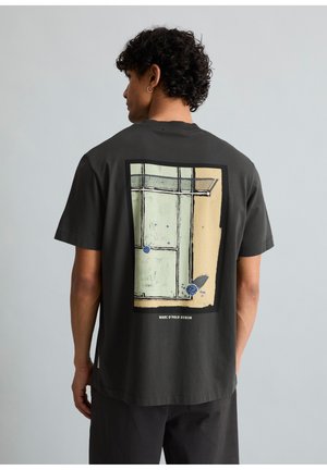 Göndör hajú férfi, fekete pólóban, amelynek hátán egy absztrakt teniszpálya és labda mintája látható, egy egyszerű szürke háttér előtt áll.