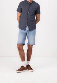 Marinblå kortärmad skjorta med vit mönster, ljusblå denimshorts, vita strumpor och bruna sneakers. Enfärgad bakgrund.