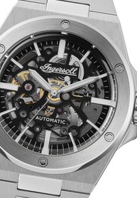 Zilveren roestvrijstalen automatische klok met een geskeletteerd zwart gezicht, witte uurmarkeringen en ingewikkelde gouden details op de beweging.