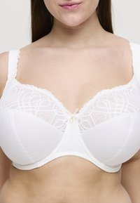Sujetador blanco con detalles de encaje en la parte superior, tela suave en las copas, bordes en forma de ondas y un pequeño acento dorado en el centro.