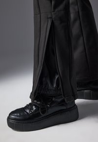 Chaussure à plateforme noire avec semelle épaisse portée avec un pantalon noir présentant une fermeture éclair ouverte révélant un poignet élastique intérieur à la cheville.