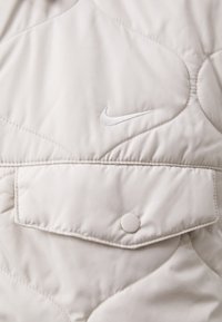 Ανοιχτό μπεζ επενδυμένο γιλέκο με λεία υφή, που διαθέτει τσέπη με κουμπί και μικρό, κεντητό λευκό λογότυπο στο στήθος.