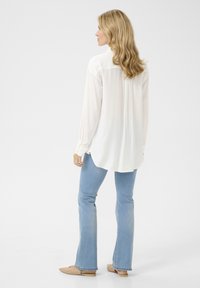 Blusa bianca a maniche lunghe con orlo leggermente curvo, abbinata a jeans chiari a zampa e ballerine beige. Tessuto morbido con vestibilità comoda.