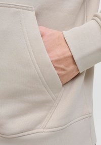 Helles beige Sweatshirt mit einer Kängurutasche. Nahaufnahme zeigt den strukturierten Stoff und sichtbare Nähdetails.