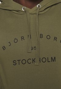 Haut à capuche vert olive avec une poche kangourou, capuche à cordon de serrage et texte imprimé noir "BJÖRN BORG 1995 STOCKHOLM" sur la poitrine.