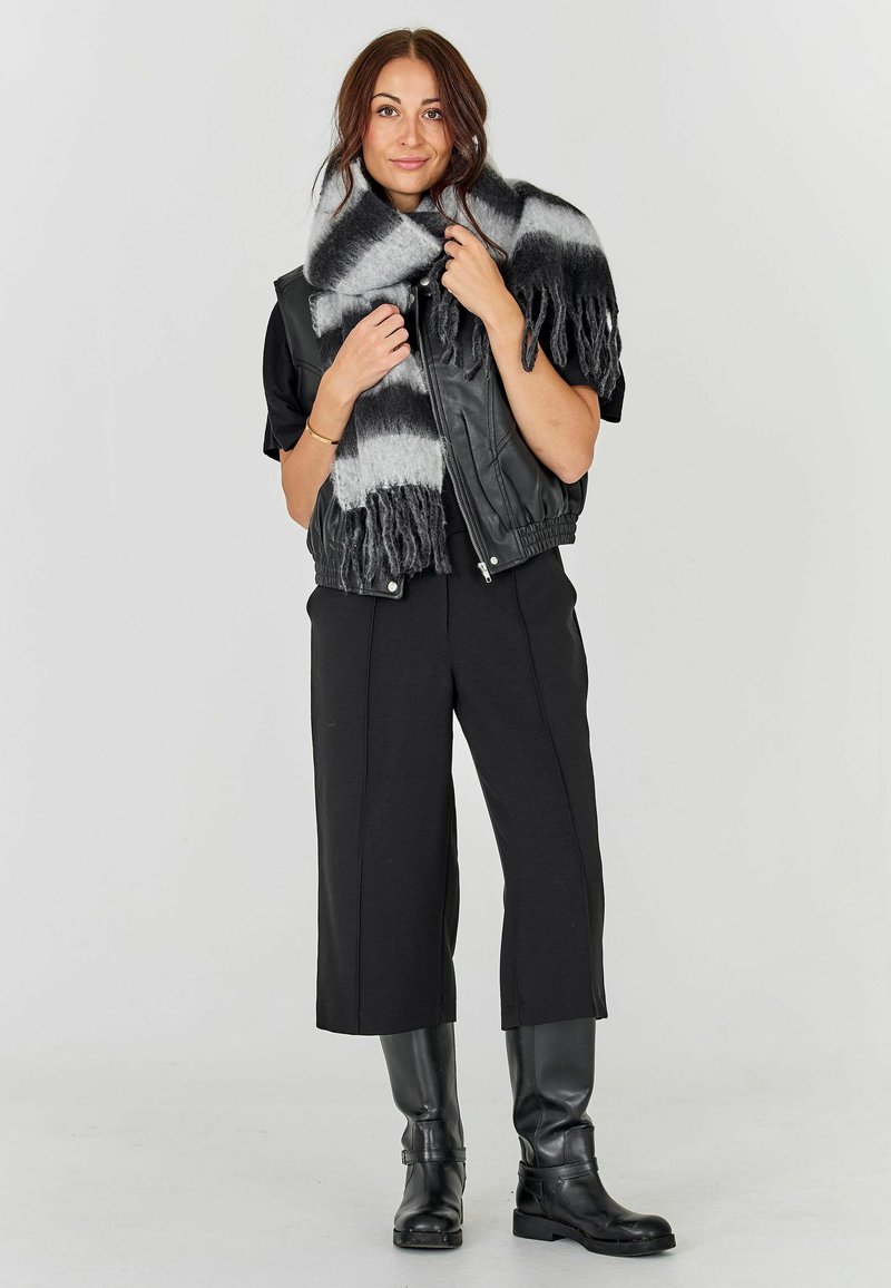 Gilet in pelle nera sopra una maglia nera, abbinato a pantaloni neri cropped e stivali neri al ginocchio. Il modello indossa una sciarpa grigia e nera a righe.