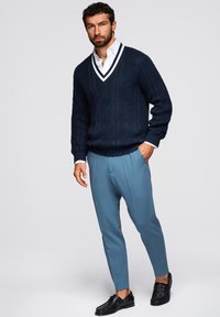 Granatowy sweter z dzianiny z białym dekoltem w serek, zestawiony z jasnoniebieskimi spodniami. Model ma na sobie czarne mokasyny; sweter ma teksturowane wzory.