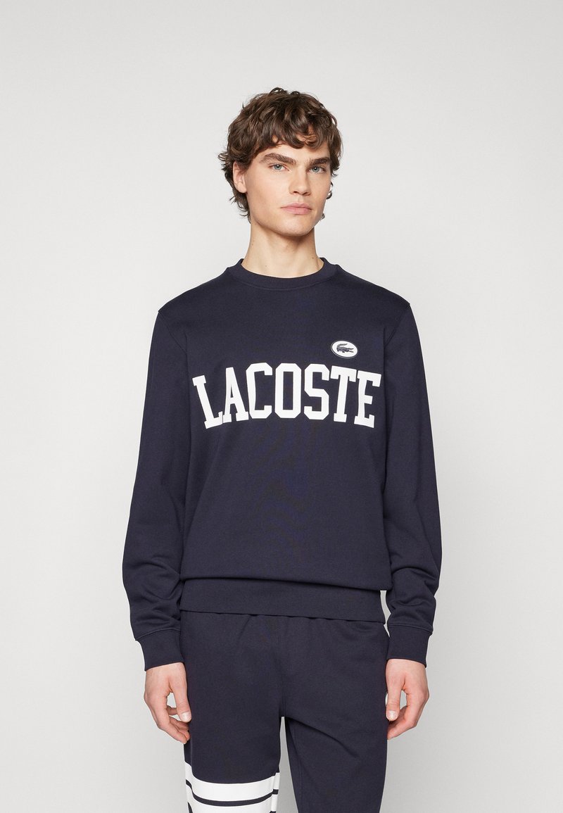 Sudadera azul marino con "LACOSTE" impreso en blanco. Cuello redondo y puños acanalados. Presenta un logo bordado en el pecho.