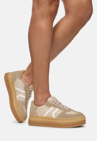 Beżowe sneakersy z białymi akcentami, z chropowatą górą z tkaniny i zamszowymi detalami. Gruba gumowa podeszwa dla dodatkowej wysokości i stabilności.