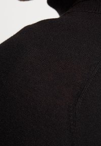 Svart tröja med en mjuk, stickad textur. Har en hög krage och en diagonal sömdetalj som visar en jämn och konsekvent tygutseende.