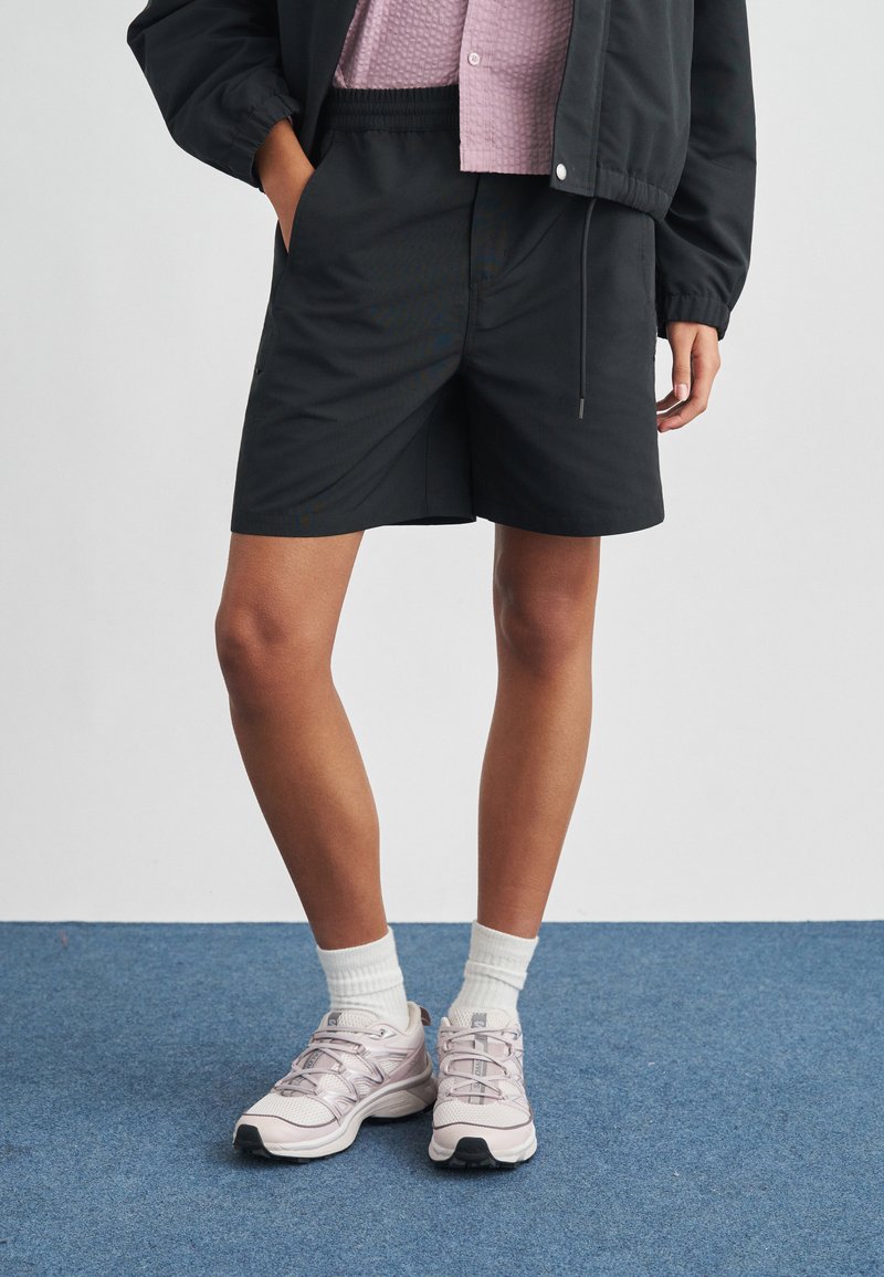 Μαύρα, ελαφριά σορτς με ελαστική μέση και πλαϊνές τσέπες, συνδυασμένα με ροζ παπούτσια για τρέξιμο και λευκές κάλτσες στη μέση σε μπλε επιφάνεια.