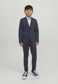 Mornarsko moder komplet s sakom z enim gumbom in elegantnimi ozkimi hlačami, kombiniran s svetlo modro srajco in belimi supergami.