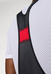 Gilet de soutien athlétique en maille noir et rouge avec une surface texturée, souligné d'une bordure noire, porté sur une chemise à manches courtes blanche.