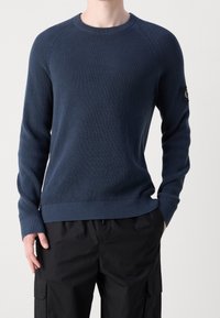 Pull en maille bleu marine avec un col ras du cou et des poignets côtelés, présentant un motif texturé et une étiquette logo sur la manche gauche.