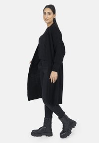 Langer schwarzer Strickcardigan über einem schwarzen Oberteil und einer Hose, kombiniert mit schwarzen Schnürkampfstiefeln; mit gerippter Textur und offenem Design.
