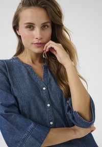 Denimskjorta i mörkblått med knappstängning fram, rund halsringning och trekvartslånga ärmar. Har en slät textur och subtil sömnad.
