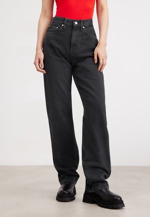 Personne portant un jean noir taille haute à jambes droites, des bottes noires épaisses et un débardeur rouge vif, debout sur un sol uni.