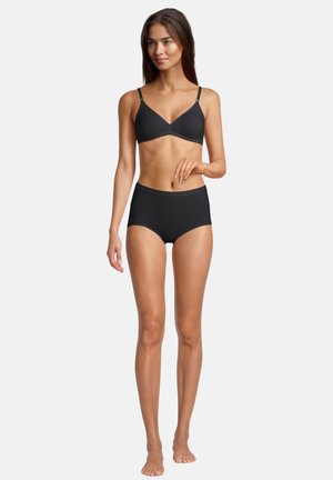 Woman standing barefoot wearing a black bra with adjustable straps and matching high-waisted briefs against white background.