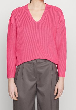 Femme portant un pull côtelé à col en V rose vif associé à un pantalon gris ample taille haute, debout devant un fond uni.