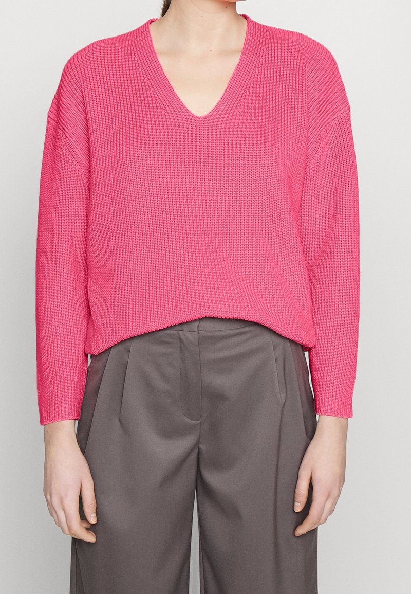 Femme portant un pull côtelé à col en V rose vif associé à un pantalon gris ample taille haute, debout devant un fond uni.