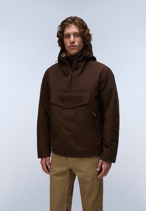 Mand med krøllet hår iført en brun Napapijri hættejakke og beige bukser mod en ensfarvet lys baggrund.