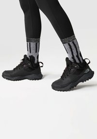 Botas de senderismo negras con suelas de goma texturizadas, punteras reforzadas y diseño de cordones. Combinadas con calcetines a rayas en blanco y negro.