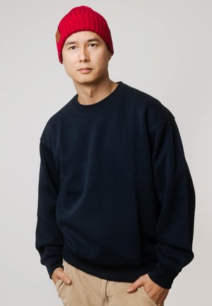 Granatowa bluza z okrągłym dekoltem i długimi rękawami. Model nosi jaskrawoczerwoną dzianinową czapkę z fakturą prążkowaną i skórzaną metką.