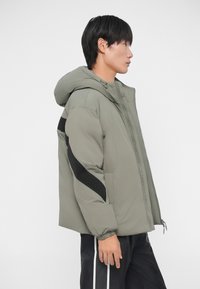 Chaqueta acolchada en verde oliva con capucha, que presenta detalles en negro y cierre de cremallera en la parte delantera. Las mangas cuentan con puños elásticos para un ajuste seguro.