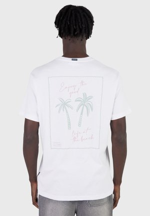 Uomo che indossa una maglietta bianca con grafica di palme e il testo "Goditi la buona vita sulla spiaggia" sul retro, in piedi contro uno sfondo uniforme.