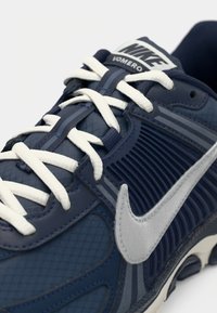 Nike Vomero sportbačiai, turintys tamsiai mėlyną viršutinę dalį, tekstūruotas tinklinis pannelis, sidabriniai akcentai, balti raišteliai ir matomas oro pado vienetas.
