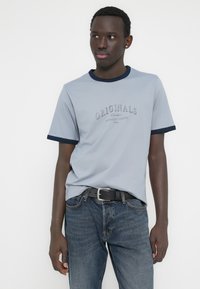 Ανοιχτό μπλε βαμβακερό μπλουζάκι με ναυτικό ριμπ γιακά και μανσέτες, με το κείμενο «ORIGINALS» σε σκούρο μπλε. Φορεμένο με σκούρο τζιν και μαύρη ζώνη.