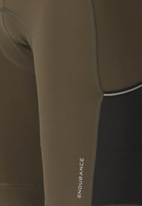 Lähikuva oliivinvihreistä ja mustista urheilushortseista, joissa on ompelusomisteita ja pystysuunnassa painettu sana "ENDURANCE".