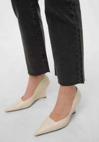 Zapatos de tacón ancho beige con punta afilada y tacón bajo, hechos de un material suave. Combinados con unos vaqueros oscuros y recortados de color gris.