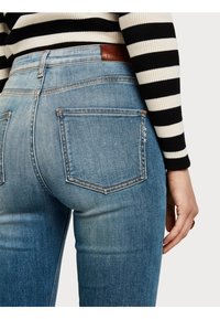 Haalaripyöreät, vaalenneet siniset farkut, joissa on korkeavyötäröinen istuvuus. Takana on kaksi taskua, joissa on ommeltu yksityiskohta sekä ruskea nahkavyötärömerkki.