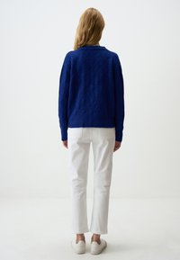 Maglione lavorato a maglia in blu profondo con motivo a rombi, abbinato a pantaloni bianchi a gamba dritta e sneaker bianche. Vista da dietro.