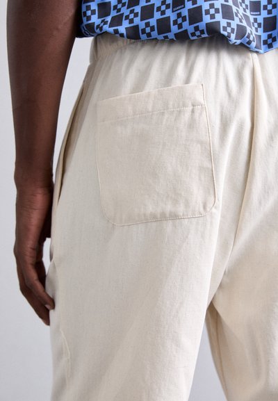 Gros plan d'une personne portant un pantalon couleur crème avec une poche arrière et une chemise à motifs bleus rentrée.
