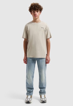 Tienerjongen met krullend haar, beige t-shirt, vervaagde blauwe spijkerbroek en witte sneakers, staand tegen een effen achtergrond.