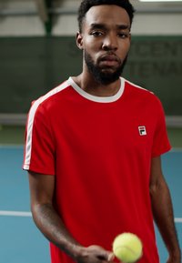 Camiseta deportiva roja con acentos blancos y logo en el pecho. Tejido suave y cuello redondo. Pelota de tenis sostenida en la mano.