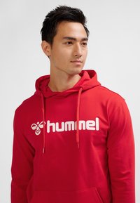 Felpa con cappuccio rossa realizzata in tessuto morbido, con un grande logo "hummel" bianco sulla parte anteriore e cordini regolabili sul cappuccio.