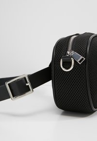 Zwarte mesh schoudertas met zilverkleurige hardware. Voorzien van een ritssluiting, een ronde metalen ring en een verstelbare schouderriem met een vierkante gesp.