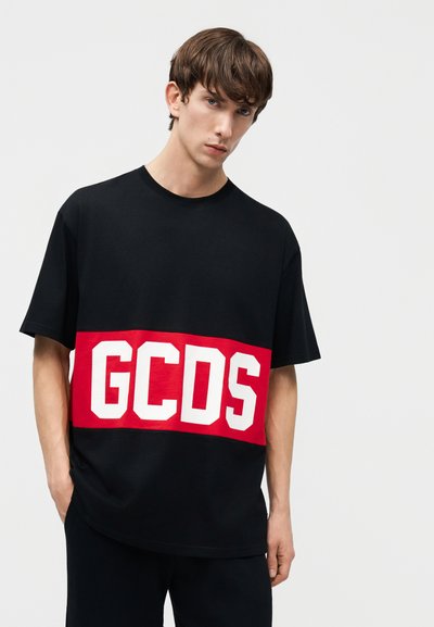 Ung mand iført en sort kortærmet t-shirt med en bred rød stribe over brystet med store hvide "GCDS" bogstaver.