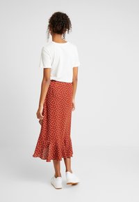 Gonna midi rossa a pois con orlo svasato, abbinata a una t-shirt bianca corta e sneaker bianche, che mostra un design casual a strati.