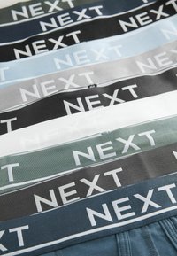 Ceintures élastiques multicolores avec "NEXT" en texte blanc. Les couleurs incluent le noir, le gris, le bleu et le vert. Texture lisse, largeur uniforme.