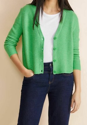 Kobieta w zielonym, teksturowanym kardiganie zapinanym na guziki, na białej koszulce i ciemnoniebieskich dżinsach, z jedną ręką w kieszeni, na jednolitym beżowym tle.