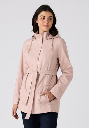 Frau trägt eine hellrosa Jacke mit Gürtel, Kapuze, goldenen Knöpfen und vorderen Taschen, steht vor einem schlichten grauen Hintergrund.