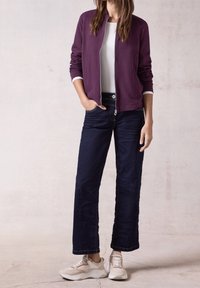 Veste bomber violette, avec des bordures blanches, associée à un jean bleu foncé ample. Baskets beige et blanches, adossé à un fond neutre.