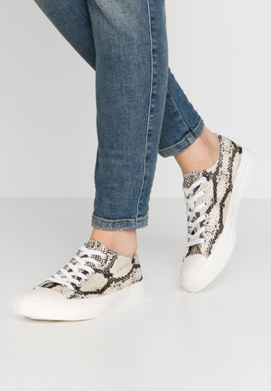 Personne portant un jean bleu et des baskets blanches avec un motif serpent noir et beige, debout sur un sol gris clair.