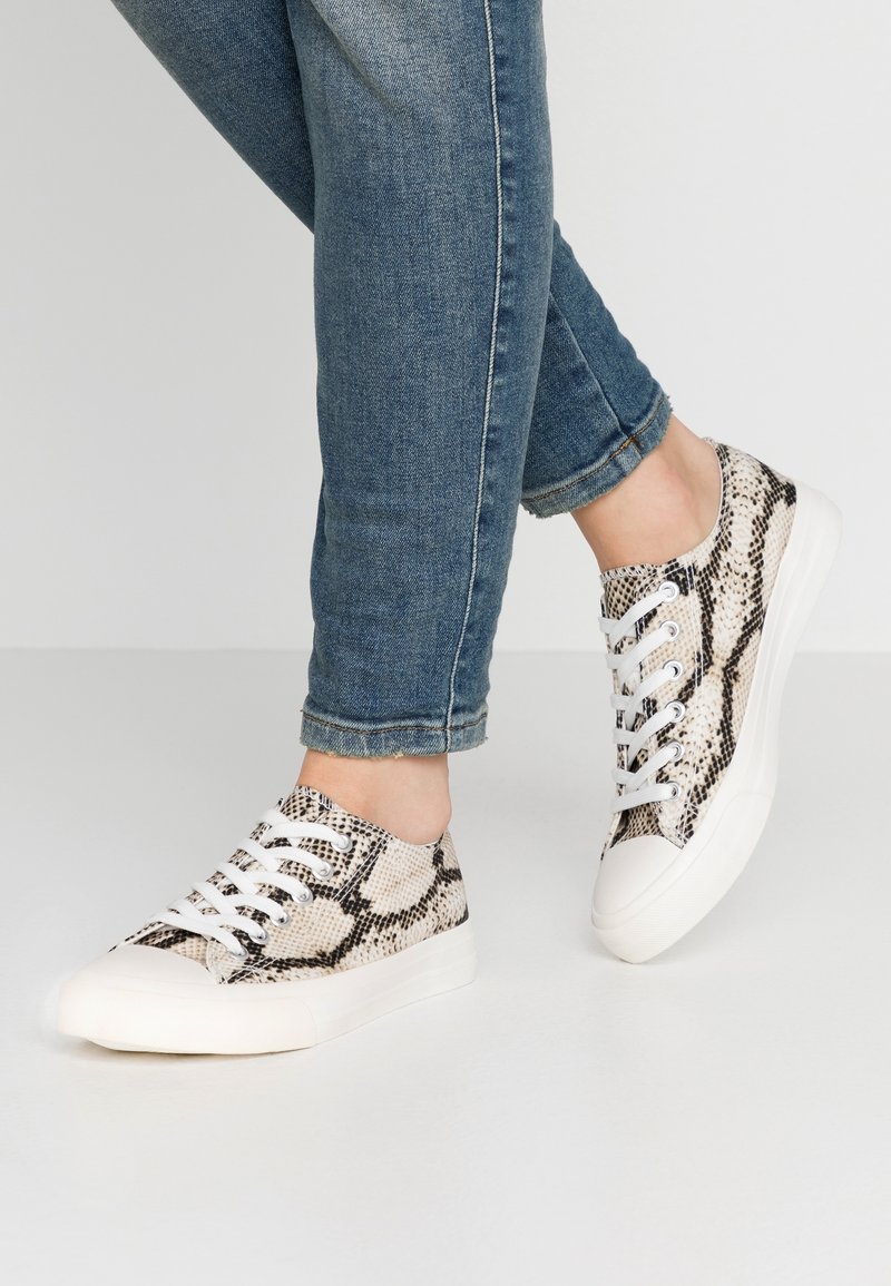 Personne portant un jean bleu et des baskets blanches avec un motif serpent noir et beige, debout sur un sol gris clair.