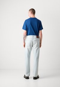 Tricou din bumbac albastru, cu mâneci scurte. Pantaloni din denimul albastru deschis, cu o petică din piele maro pe spate. Pantofi negri. Fundal alb simplu.
